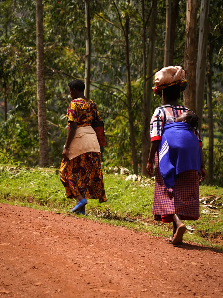 Two women walking a red-earth road, one carrying a bundle on her head and a baby on her back
