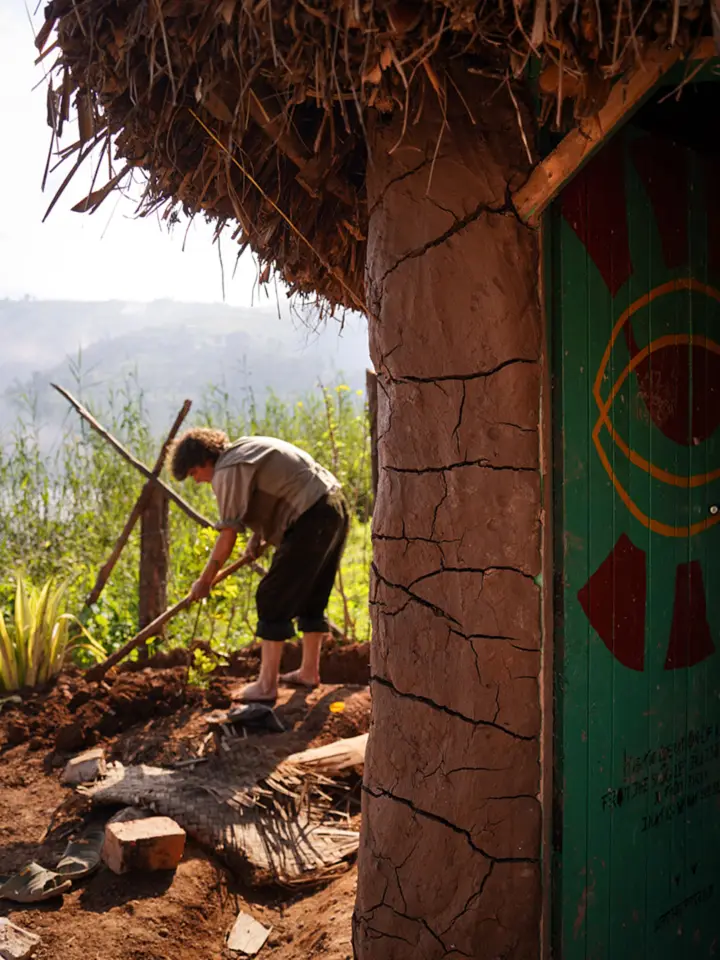A man digging the ground beside a thatched hut with a painted green door